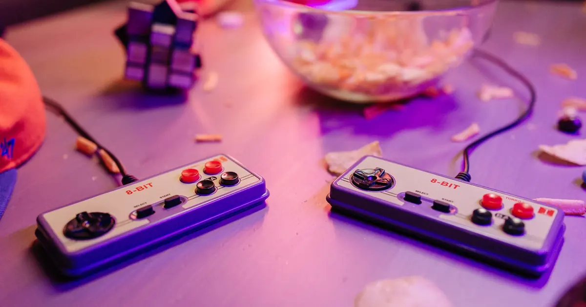 Two grey retro-style game controllers with red buttons on a table under purple light, a snack bowl behind them
